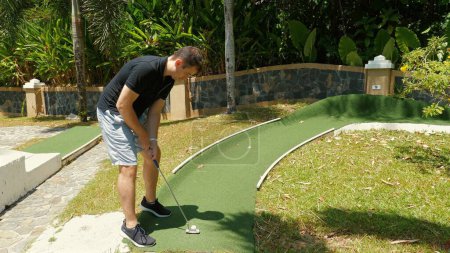 Téléchargez les photos : Homme adulte actif jouant au mini-golf par une journée ensoleillée, profitant d'activités de loisirs de plein air sur putting green bien entretenu. Sports de plein air et divertissement. - en image libre de droit