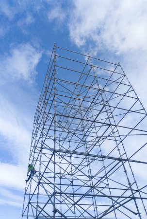 Foto de Trabajador no identificado en altura desmantelando una torre de andamio. Cielo nublado azul sobre fondo - Imagen libre de derechos