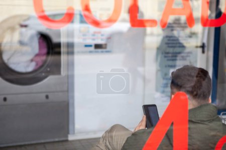 Photo for Young man waiting with smartphone at self-service laundry. Vview through the glass - Royalty Free Image