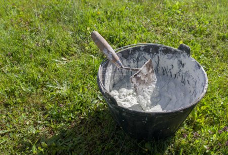 Photo for Bucket full white adhesive mortar ready for applicate. Green grass as background - Royalty Free Image