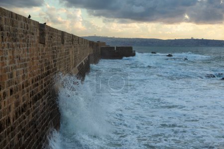 Foto de Las olas del mar golpearon la muralla de la fortaleza del Acre antiguo contra el fondo de un cielo con nubes de trueno - Imagen libre de derechos