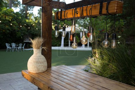 Téléchargez les photos : Ampoules décoratives suspendues sur une barre en bois et épis de blé dans une cruche sur fond de pelouse de mariage - en image libre de droit