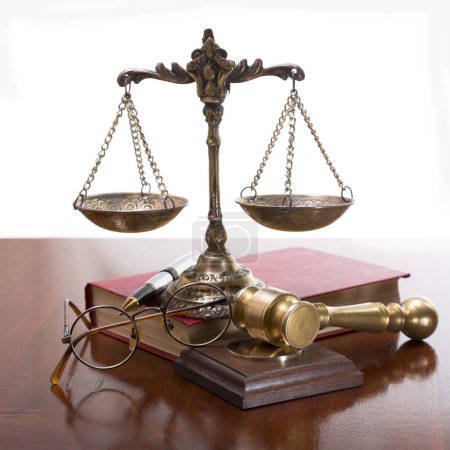 Foto de Escamas de bronce y un mazo de juez, gafas de marco redondo, un bolígrafo y un libro con una cubierta roja sobre una mesa de madera barnizada - Imagen libre de derechos