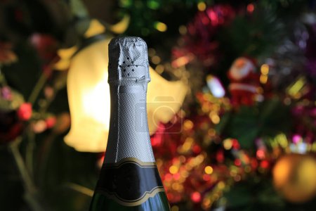 Foto de A bottle of closed champagne against the backdrop of a glowing Christmas tree with toys and garlands - Imagen libre de derechos