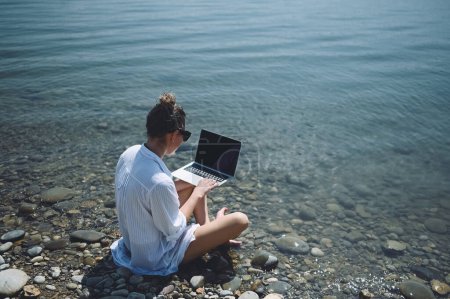 Téléchargez les photos : Jeune femme voyageur pigiste portant chemise blanche n'importe où travaillant en ligne à l'extérieur en utilisant un ordinateur portable bénéficiant d'une vue sur le lac. Heureux downshifter féminin dans les lunettes de soleil tenant ordinateur à la côte de la mer - en image libre de droit