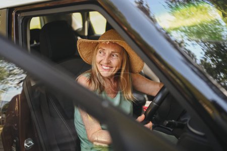 Téléchargez les photos : Conductrice senior heureuse en chapeau de paille conduisant assise dans une nouvelle voiture, souriant en regardant la caméra profiter du voyage. Cours de conduite et assurance vie. Activité des retraités et concept de road trip - en image libre de droit