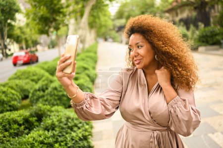 Attractive happy stylish plus size African American woman student freelancer afro hair making selfie with smartphone outside. Young lady in silk dress in big urban city street at summer. Diversity. 