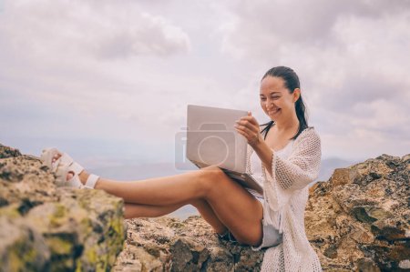 Téléchargez les photos : Jeune femme voyageur indépendant travaillant en ligne n'importe où à l'extérieur en utilisant un ordinateur portable bénéficiant d'une vue sur la montagne. Happy downshifter femme tenant l'ordinateur dans un lieu de travail de vue incroyable inhabituel à l'été - en image libre de droit