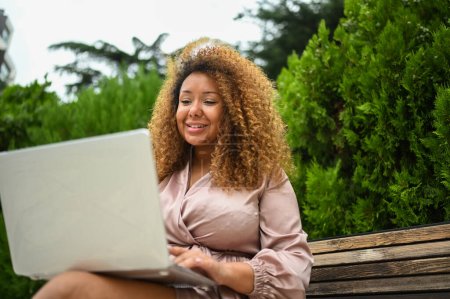 Téléchargez les photos : Attrayant heureux élégant plus la taille afro-américaine étudiante freelancer afro cheveux étudiant en ligne travaillant sur ordinateur portable à l'extérieur au parc d'été. La diversité. Travail à distance, enseignement à distance - en image libre de droit