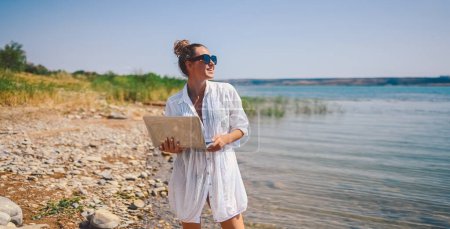 Téléchargez les photos : Jeune femme voyageur pigiste portant chemise blanche n'importe où travaillant en ligne à l'extérieur en utilisant un ordinateur portable bénéficiant d'une vue sur le lac. Heureux downshifter féminin dans les lunettes de soleil tenant ordinateur à la côte de la mer - en image libre de droit