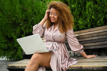 Foto de Atractivo feliz elegante más tamaño mujer afroamericana estudiante freelancer afro pelo estudiando en línea trabajando en el ordenador portátil fuera en el parque de verano. Diversidad. Trabajo remoto, educación a distancia - Imagen libre de derechos