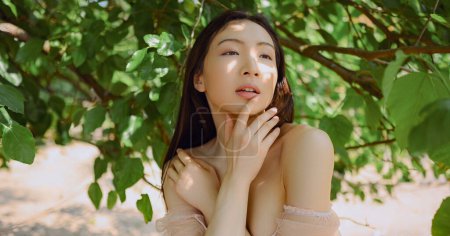 Nahaufnahme Außenporträt Schöne junge chinesische asiatische Frau perfekte Haut posiert außerhalb sonnigen Sommertages grünes Laub. Natürliche Gesichtsbehandlung. Kosmetologie, Hautpflege und Wellness. Fragmentierungskonzept