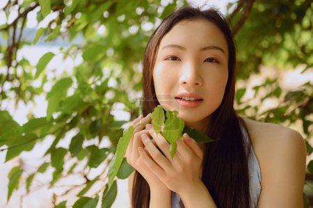 Close up outdoor portrait Beautiful young Chinese Asian woman perfect skin posing outside sunny summer day green foliage. Natural facial treatment. Cosmetology, skin care and spa. Fragnance concept