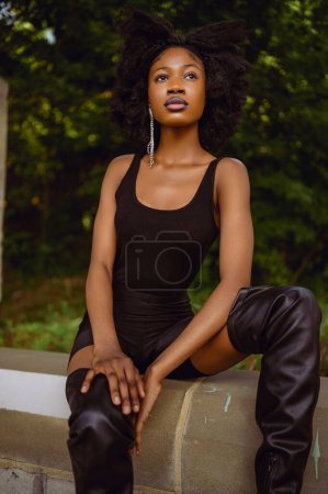 Mode im Freien Streetstyle Porträt Schöne junge Afroamerikanerin posiert draußen auf städtischen Stadtlandschaft Sommertag trägt insgesamt schwarze hohe Lederstiefel.