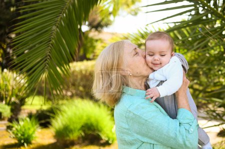 Beautiful happy smiling senior elderly woman holding on hands cute little baby boy. Grandmother and grandson having fun time together at tropical summer day in park. Multigenerational family concept