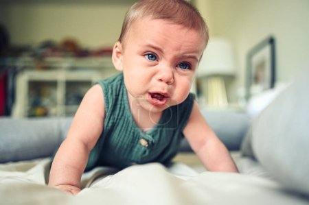 Foto de Primer plano retrato de niño pequeño niño lindo divertido niño rubio con ojos azules en traje de cuerpo de lino verde llorando en el interior de la habitación de la guardería. Rabieta infantil. Dificultad para acostar al bebé - Imagen libre de derechos