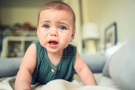 Foto de Primer plano retrato de niño pequeño niño lindo divertido niño rubio con ojos azules en traje de cuerpo de lino verde llorando en el interior de la habitación de la guardería. Rabieta infantil. Dificultad para acostar al bebé - Imagen libre de derechos