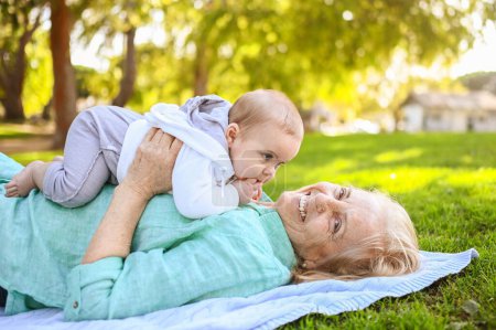 Hermosa mujer mayor sonriente feliz anciana acostada en la hierba con lindo niño pequeño. La abuela y el nieto se divierten juntos en el parque tropical de verano. Concepto familiar multigeneracional