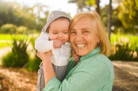 Hermosa feliz sonriente anciana mayor sosteniendo en las manos lindo niño pequeño. La abuela y el nieto se divierten juntos en el día tropical de verano en el parque. Concepto familiar multigeneracional