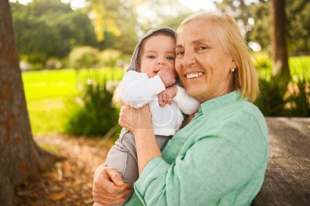 Schöne glücklich lächelnde Seniorin, die an den Händen einen niedlichen kleinen Jungen hält. Großmutter und Enkel amüsieren sich gemeinsam an einem tropischen Sommertag im Park. Familienkonzept für mehrere Generationen