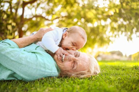 Schöne, glücklich lächelnde Seniorin, die mit einem niedlichen kleinen Jungen im Gras liegt. Großmutter und Enkel amüsieren sich gemeinsam im tropischen Sommertagspark. Familienkonzept für mehrere Generationen