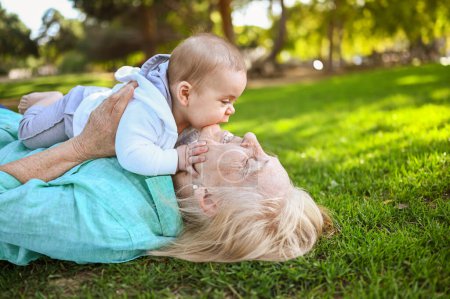 Beautiful happy smiling senior elderly woman lying on grass with cute little baby boy. Grandmother and grandson having fun time together at tropical summer day park. Multigenerational family concept