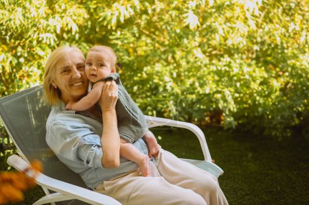 Hermosa feliz sonriente anciana mayor sosteniendo en las manos lindo bebé niño sentado al aire libre. Abuela nieto divertirse juntos en el soleado día de verano en el jardín o el parque