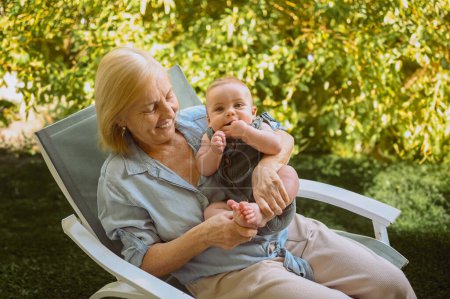 Schöne glücklich lächelnde ältere Frau, die an den Händen den niedlichen kleinen Jungen hält, der draußen sitzt. Großmutter-Enkel beim gemeinsamen Spaß an sonnigen Sommertagen im Garten oder Park
