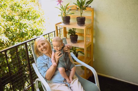 Schöne glücklich lächelnde ältere Frau, die an den Händen den niedlichen kleinen Jungen hält, der auf einem Schaukelstuhl im Freien sitzt. Großmutter-Enkel amüsiert sich bei sonnigem Sommertag auf Balkonterrasse