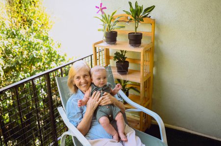 Schöne glücklich lächelnde ältere Frau, die an den Händen den niedlichen kleinen Jungen hält, der auf einem Schaukelstuhl im Freien sitzt. Großmutter-Enkel amüsiert sich bei sonnigem Sommertag auf Balkonterrasse
