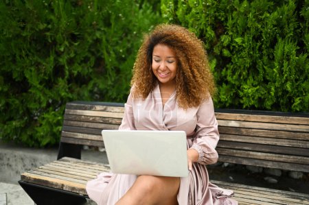 Attractive happy stylish plus size African American woman student freelancer afro hair studying online working on laptop computer outside at summer park. Diversity. Remote work, distance education