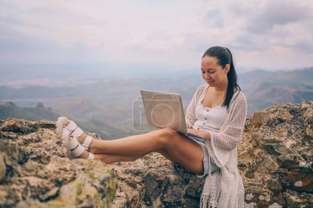 Téléchargez les photos : Jeune femme voyageur indépendant travaillant en ligne n'importe où à l'extérieur en utilisant un ordinateur portable bénéficiant d'une vue sur la montagne. Happy downshifter femme tenant l'ordinateur dans un lieu de travail de vue incroyable inhabituel à l'été - en image libre de droit