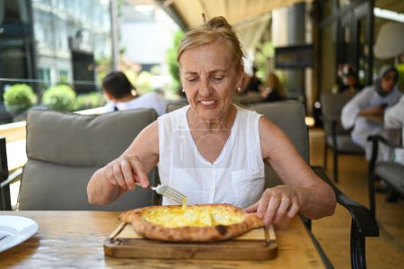 Happy senior woman with perfect smile eating adjarian khachapuri outdoors at summer sidewalk street cafe or restaurant terrace. Retired mature people holiday vacation, active lifestyle concept