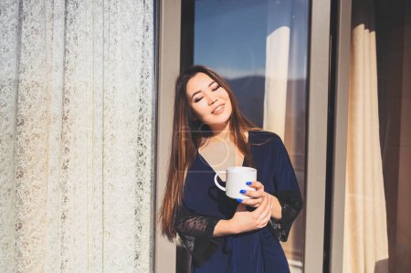 Hermosa mujer asiática sonriente disfrutando de café o té en el balcón soleado. Joven dama feliz en albornoz azul relajante tomar el sol en la terraza en verano