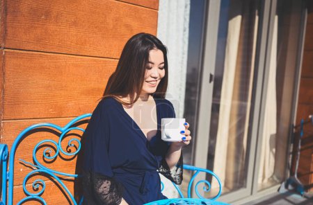 Hermosa mujer asiática sonriente disfrutando de café o té en el balcón soleado. Joven dama feliz en albornoz azul relajante tomar el sol en la terraza en verano