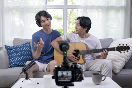 Photo for Young Asian gay couple blogger vlogger and online influencer recording musical video content playing guitar and singing at home. LGBT male couple performing and shooting clip for social media. - Royalty Free Image