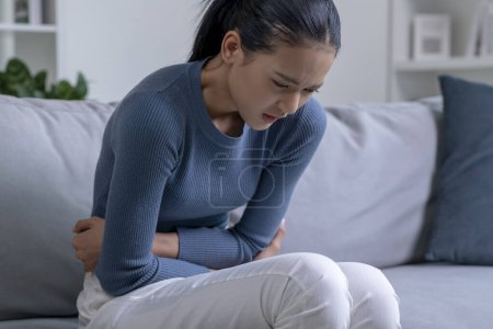 Mujer asiática joven que sufre de fuerte dolor abdominal mientras está sentada en el sofá en casa. Mujer enferma con dolor de estómago.