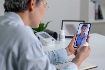 Asiatin mit Smartphone während einer Online-Beratung mit ihrem Arzt im Wohnzimmer, telemedizinisches Konzept