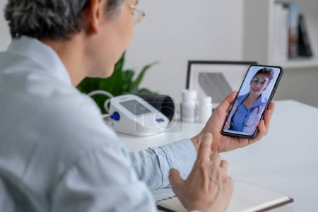 Photo for Asian woman with smartphone during an online consultation with her doctor in her living room, telemedicine concept - Royalty Free Image
