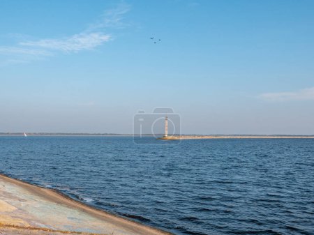 Téléchargez les photos : Vieux phare en béton sur le bord de la mer de Kiev crache avec des oiseaux volant au-dessus, eau bleue calme avec un yacht blanc, et ciel bleu avec des nuages de plumes, étroite bande de rive à l'horizon. - en image libre de droit