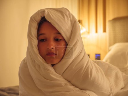 Cute teen girl lying on a bed cocooned comfortably in a puffy white blanket with soft yellow light at the background in the evening. Relaxed resting mood concept.