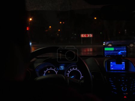 Driver sits behind the wheel and drives in a car through dark city at night. Raindrops on a windshield, highlighted dashboard, road traffic with blurry car lights, and street lamps.