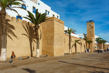 Téléchargez les photos : Maroc. Essaouira. Les gens marchant le long des remparts et de la tour de l'horloge - en image libre de droit
