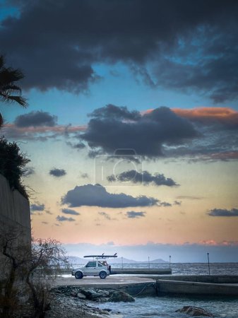 Photo for Mid distance view of off-road vehicle with paddleboard on roof parked at beach against seascape and cloudy sky during sunset - Royalty Free Image