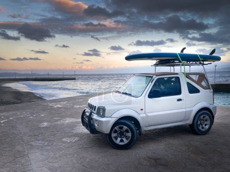 Foto de Vehículo todoterreno con remo en el techo estacionado en la playa contra el paisaje marino y el cielo nublado al atardecer - Imagen libre de derechos