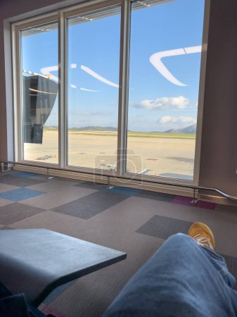 Foto de Sección baja del hombre con pantalones vaqueros azules sentado en la sala de espera frente a la pista vista a través de la ventana del aeropuerto - Imagen libre de derechos
