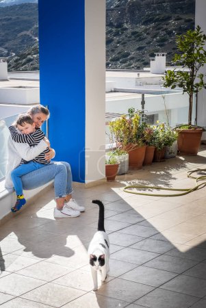 Foto de Amar caucásico mediana edad madre abrazando hijo y mirando gato mientras sentado en balcón durante día soleado - Imagen libre de derechos