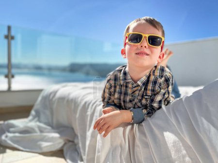 Foto de Retrato de niño lindo con gafas de sol y descansando en la cama contra el cielo azul claro en el patio durante el día soleado - Imagen libre de derechos