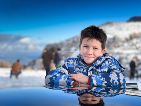 Foto de Retrato del niño sonriente con abrigo de invierno con los brazos cruzados apoyados en el techo del coche contra la montaña y el cielo despejado - Imagen libre de derechos