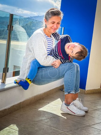 Foto de Amorosa madre caucásica mirando lindo hijo cerrando los ojos y haciendo cara mientras sentado en su regazo en balcón - Imagen libre de derechos